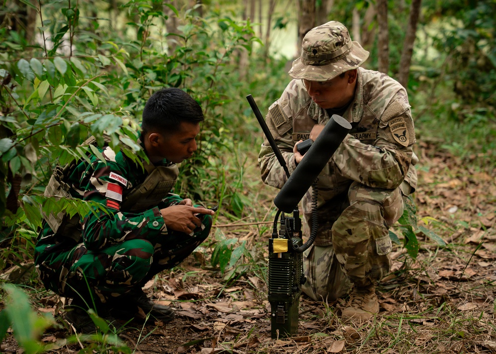 Two military personnel are crouched in a forested area examining a piece of communications or electronic equipment placed on the ground. One soldier wears Indonesian Army (TNI-AD) camouflage with an 'INDONESIA' patch and red-white flag, while the other wears US Army MultiCam uniform with an 'AIRBORNE' tab and Arctic patch, identified by a 'PANT-' name tape. This appears to be a joint military training exercise between Indonesian and US Army forces.