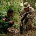Two military personnel are crouched in a forested area examining a piece of communications or electronic equipment placed on the ground. One soldier wears Indonesian Army (TNI-AD) camouflage with an 'INDONESIA' patch and red-white flag, while the other wears US Army MultiCam uniform with an 'AIRBORNE' tab and Arctic patch, identified by a 'PANT-' name tape. This appears to be a joint military training exercise between Indonesian and US Army forces.
