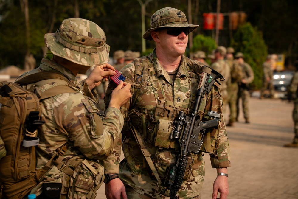 Two U.S. Army soldiers in MultiCam (OCP) uniforms are shown in the foreground, with one soldier affixing a U.S. flag patch to the other's shoulder strap. The soldier on the right is armed with an M4-style carbine and wears Staff Sergeant rank insignia. Additional soldiers are visible in the background, suggesting a formation or pre-mission assembly.