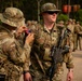 Two U.S. Army soldiers in MultiCam (OCP) uniforms are shown in the foreground, with one soldier affixing a U.S. flag patch to the other's shoulder strap. The soldier on the right is armed with an M4-style carbine and wears Staff Sergeant rank insignia. Additional soldiers are visible in the background, suggesting a formation or pre-mission assembly.