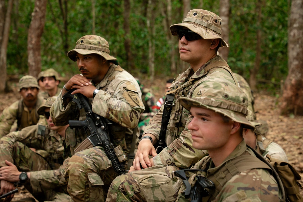 A group of military personnel in MultiCam/OCP camouflage uniforms are seated in a wooded area, appearing to receive a briefing or instruction. The soldiers are equipped with weapons, tactical gear, and boonie hats. The setting suggests a field training exercise or operational pause in a tropical or subtropical forest environment.