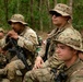 A group of military personnel in MultiCam/OCP camouflage uniforms are seated in a wooded area, appearing to receive a briefing or instruction. The soldiers are equipped with weapons, tactical gear, and boonie hats. The setting suggests a field training exercise or operational pause in a tropical or subtropical forest environment.