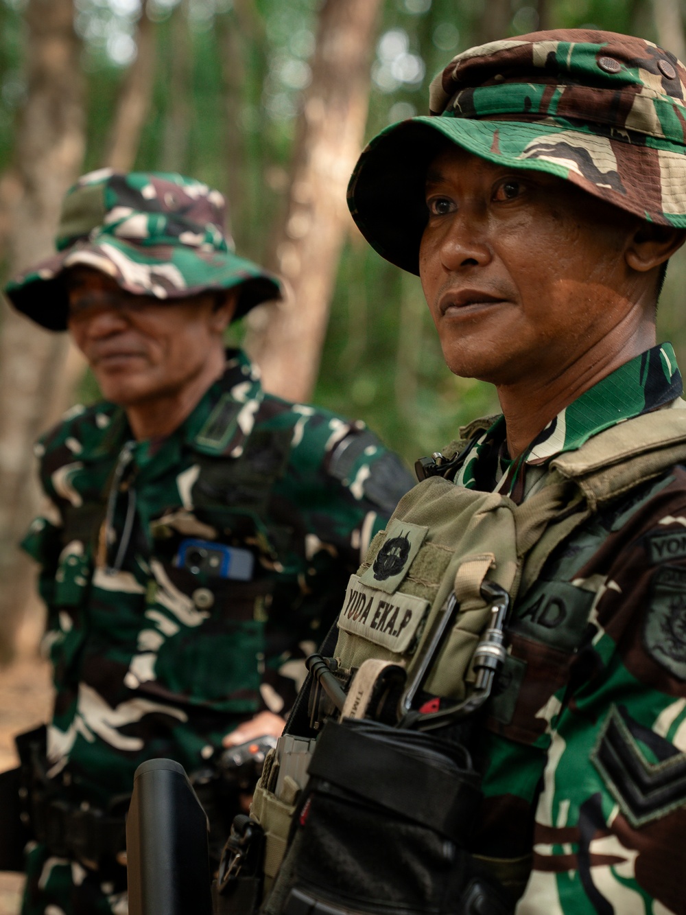 Two military personnel in a forested outdoor setting, wearing Indonesian Army (TNI-AD) camouflage uniforms and boonie hats. The foreground soldier is equipped with tactical gear including a plate carrier vest with a name tape reading 'YUDA EKA.P' and carries a weapon. Both soldiers appear to be standing at attention or observing something off-frame.