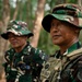 Two military personnel in a forested outdoor setting, wearing Indonesian Army (TNI-AD) camouflage uniforms and boonie hats. The foreground soldier is equipped with tactical gear including a plate carrier vest with a name tape reading 'YUDA EKA.P' and carries a weapon. Both soldiers appear to be standing at attention or observing something off-frame.