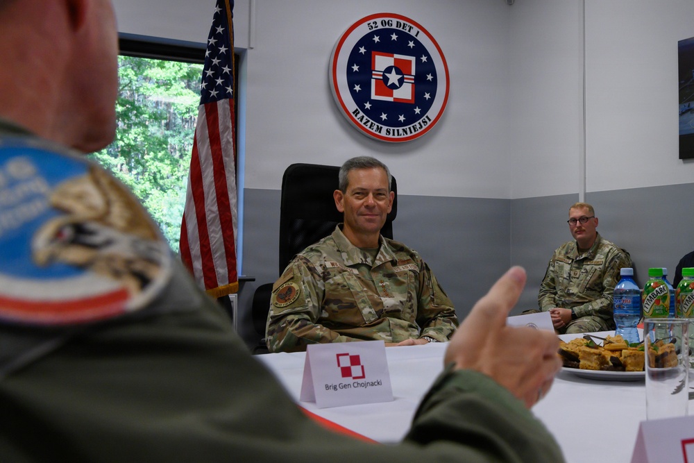 A military meeting or briefing is taking place at a facility associated with the 52nd Operations Group Detachment 1, as indicated by the unit insignia on the wall. A name placard identifies 'Brig Gen Chojnacki' at the conference table, suggesting a bilateral or multinational military engagement. An American flag is visible in the background alongside the unit patch bearing the motto 'Razem Silniejsi' (Polish for 'Stronger Together').
