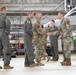 Military personnel are gathered inside an aircraft hangar, with two individuals in the foreground engaged in a handshake. The group includes personnel wearing both flight suits and camouflage (OCP/MultiCam) uniforms. Fighter aircraft, likely F-16s, are visible in the background along with weapons and equipment.