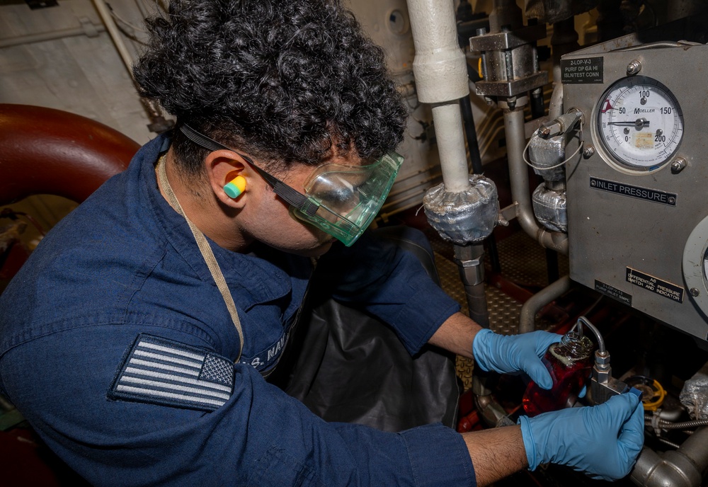 A U.S. Navy sailor in a dark blue uniform is performing maintenance work on mechanical equipment aboard a naval vessel. The sailor is wearing personal protective equipment including green safety goggles, blue nitrile gloves, and foam ear plugs while working on what appears to be a pressurized fluid system. A pressure gauge labeled 'INLET PRESSURE' with a scale of 0-200 is visible on the equipment panel.