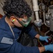 A U.S. Navy sailor in a dark blue uniform is performing maintenance work on mechanical equipment aboard a naval vessel. The sailor is wearing personal protective equipment including green safety goggles, blue nitrile gloves, and foam ear plugs while working on what appears to be a pressurized fluid system. A pressure gauge labeled 'INLET PRESSURE' with a scale of 0-200 is visible on the equipment panel.