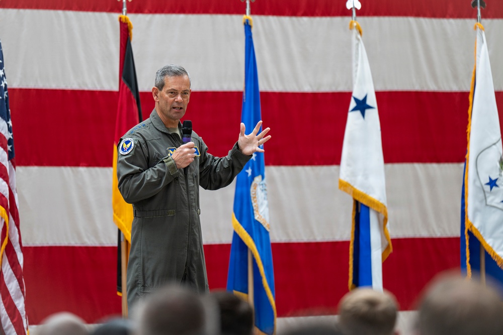 A military officer in an olive green flight suit is addressing an audience while holding a microphone, gesturing with his right hand. The setting appears to be an official military ceremony or briefing held in front of a large American flag backdrop. Several military flags and guidons are visible in the background, including what appears to be a two-star general officer flag.