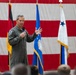 A military officer in an olive green flight suit is addressing an audience while holding a microphone, gesturing with his right hand. The setting appears to be an official military ceremony or briefing held in front of a large American flag backdrop. Several military flags and guidons are visible in the background, including what appears to be a two-star general officer flag.