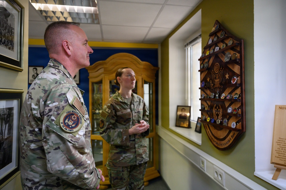 Two U.S. Air Force personnel in OCP (Operational Camouflage Pattern) uniforms are examining a military challenge coin display rack mounted on a wall. The male service member wears a 'Chief Master Sergeant of the Air Force' patch on his shoulder, indicating a high-ranking enlisted position. They appear to be in a heritage or memorabilia room containing framed photographs and military artifacts.