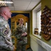 Two U.S. Air Force personnel in OCP (Operational Camouflage Pattern) uniforms are examining a military challenge coin display rack mounted on a wall. The male service member wears a 'Chief Master Sergeant of the Air Force' patch on his shoulder, indicating a high-ranking enlisted position. They appear to be in a heritage or memorabilia room containing framed photographs and military artifacts.