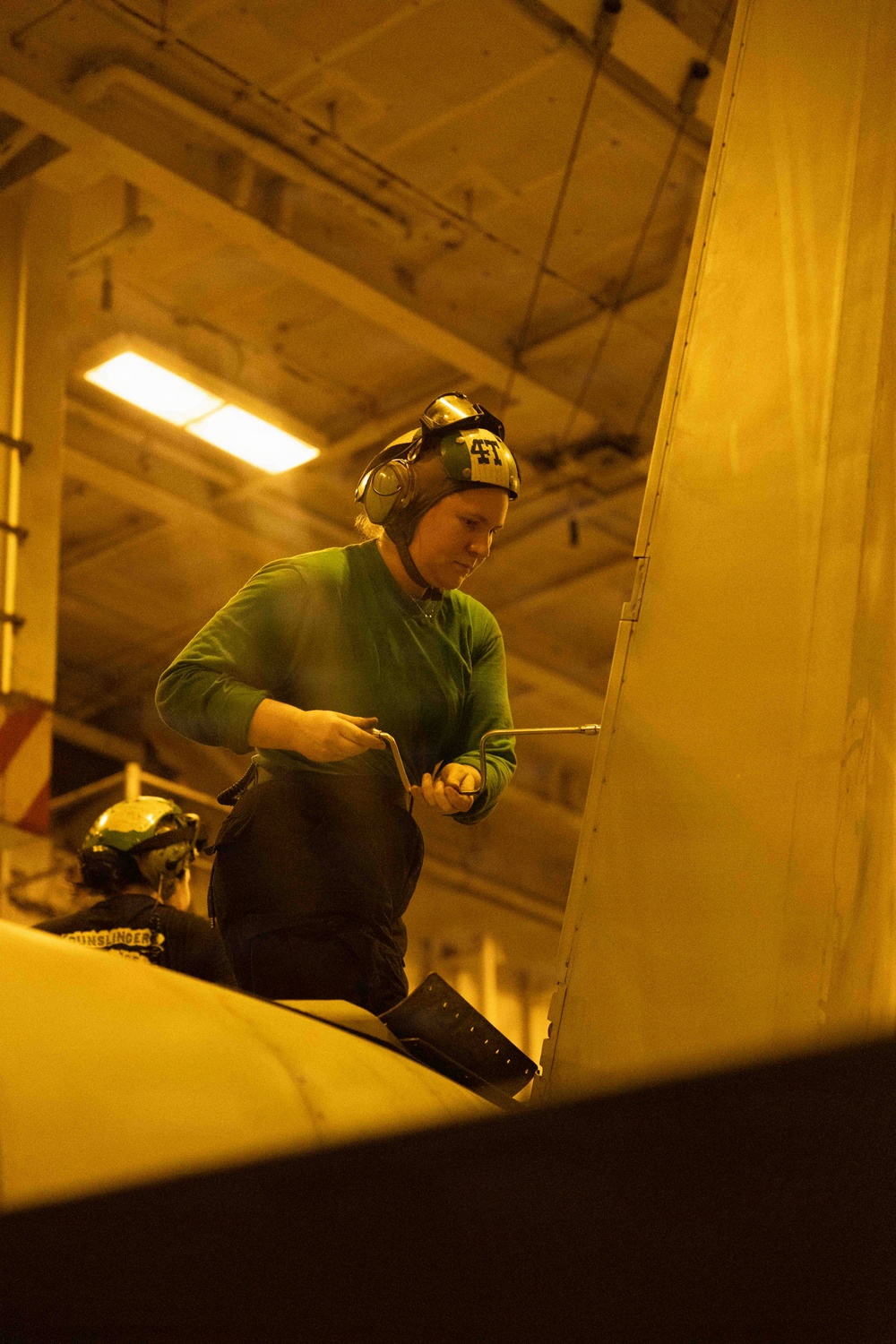 A military aviation technician wearing a green jersey and helmet with ear protection (numbered '41') performs maintenance work on an aircraft in what appears to be a naval vessel's hangar bay. The warm yellow lighting is characteristic of a ship's interior hangar deck. A second crew member wearing a helmet marked 'GUNSLINGER' is partially visible in the lower left.