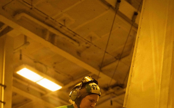 A military aviation technician wearing a green jersey and helmet with ear protection (numbered '41') performs maintenance work on an aircraft in what appears to be a naval vessel's hangar bay. The warm yellow lighting is characteristic of a ship's interior hangar deck. A second crew member wearing a helmet marked 'GUNSLINGER' is partially visible in the lower left.