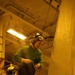A military aviation technician wearing a green jersey and helmet with ear protection (numbered '41') performs maintenance work on an aircraft in what appears to be a naval vessel's hangar bay. The warm yellow lighting is characteristic of a ship's interior hangar deck. A second crew member wearing a helmet marked 'GUNSLINGER' is partially visible in the lower left.