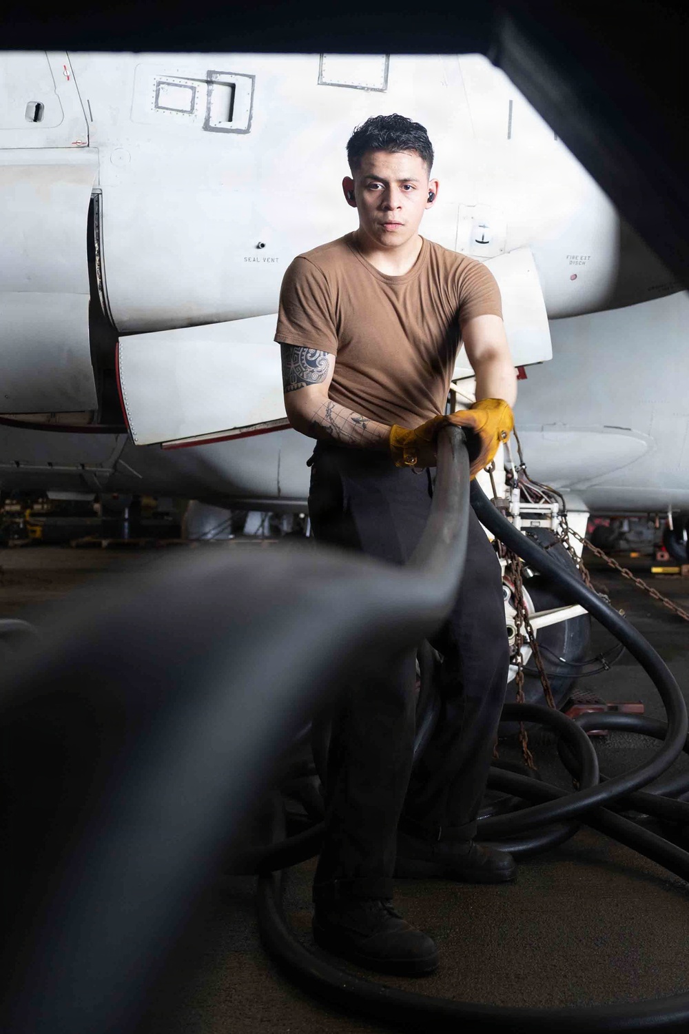 A young military or aviation maintenance personnel is working in what appears to be an aircraft hangar, handling large hoses or cables near a white military aircraft. The subject is wearing a tan/brown t-shirt, dark trousers, and yellow work gloves, with visible tattoos on his right arm. The image is a professional portrait-style photograph taken in a low-light hangar environment.