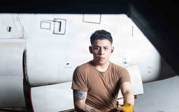 A young military or aviation maintenance personnel is working in what appears to be an aircraft hangar, handling large hoses or cables near a white military aircraft. The subject is wearing a tan/brown t-shirt, dark trousers, and yellow work gloves, with visible tattoos on his right arm. The image is a professional portrait-style photograph taken in a low-light hangar environment.