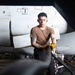 A young military or aviation maintenance personnel is working in what appears to be an aircraft hangar, handling large hoses or cables near a white military aircraft. The subject is wearing a tan/brown t-shirt, dark trousers, and yellow work gloves, with visible tattoos on his right arm. The image is a professional portrait-style photograph taken in a low-light hangar environment.
