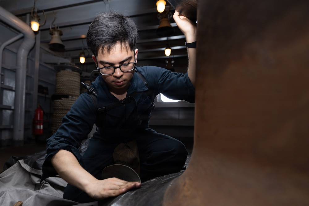 A young man wearing glasses and a dark navy coverall uniform is crouched down inspecting or working on a large metallic object in what appears to be the interior of a naval vessel or historic ship. He is using one hand to hold a light source above while the other hand examines the surface below. The environment shows industrial ship interior features including overhead lights, coiled rope, pipes, and a fire extinguisher.