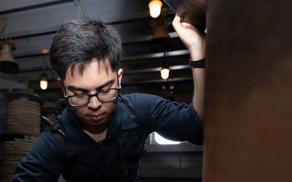 A young man wearing glasses and a dark navy coverall uniform is crouched down inspecting or working on a large metallic object in what appears to be the interior of a naval vessel or historic ship. He is using one hand to hold a light source above while the other hand examines the surface below. The environment shows industrial ship interior features including overhead lights, coiled rope, pipes, and a fire extinguisher.