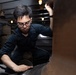 A young man wearing glasses and a dark navy coverall uniform is crouched down inspecting or working on a large metallic object in what appears to be the interior of a naval vessel or historic ship. He is using one hand to hold a light source above while the other hand examines the surface below. The environment shows industrial ship interior features including overhead lights, coiled rope, pipes, and a fire extinguisher.