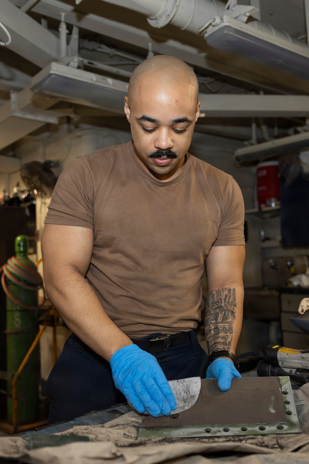 A bald man with a mustache and forearm tattoo is working in what appears to be a military ship's workshop or maintenance space. He is wearing a brown t-shirt, dark pants, and blue nitrile gloves while sanding or polishing a metal panel. The environment features overhead pipes, cables, and industrial equipment consistent with a naval vessel's interior.