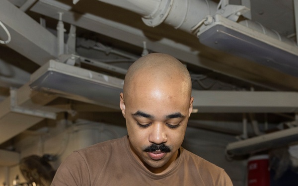A bald man with a mustache and forearm tattoo is working in what appears to be a military ship's workshop or maintenance space. He is wearing a brown t-shirt, dark pants, and blue nitrile gloves while sanding or polishing a metal panel. The environment features overhead pipes, cables, and industrial equipment consistent with a naval vessel's interior.
