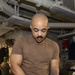 A bald man with a mustache and forearm tattoo is working in what appears to be a military ship's workshop or maintenance space. He is wearing a brown t-shirt, dark pants, and blue nitrile gloves while sanding or polishing a metal panel. The environment features overhead pipes, cables, and industrial equipment consistent with a naval vessel's interior.