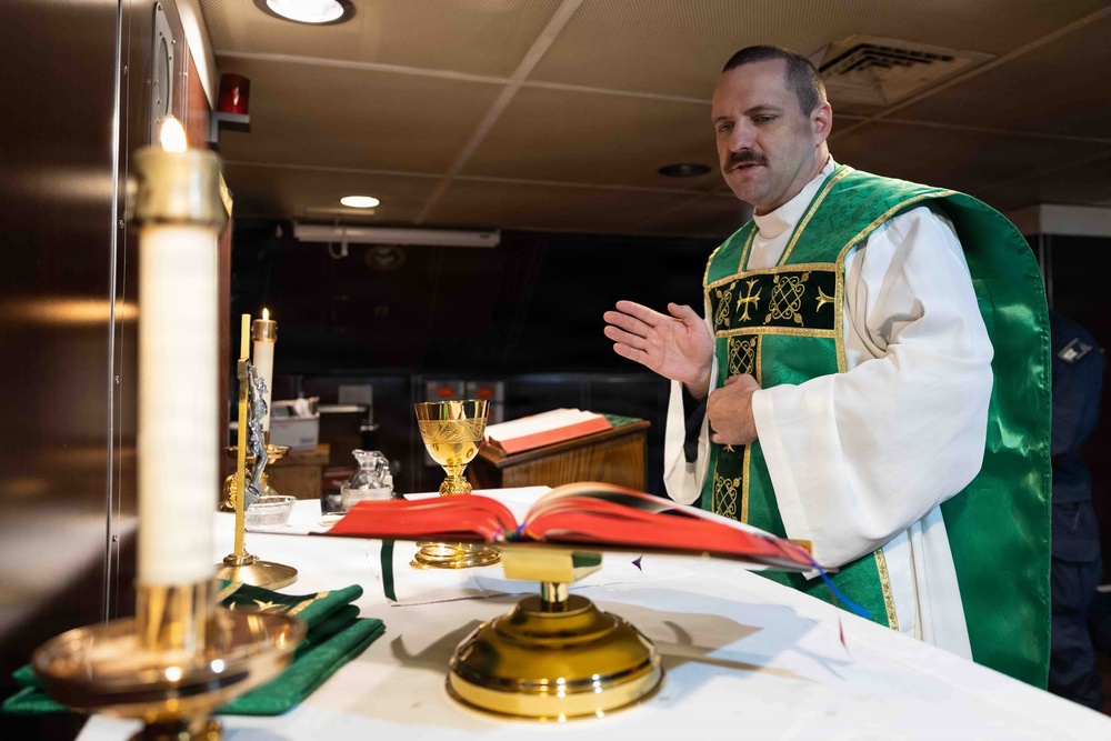 A Catholic priest wearing green liturgical vestments (chasuble) over a white alb is celebrating Mass at an altar. The altar is set with traditional Catholic Mass items including a chalice, candles, and an open missal. The setting appears to be a non-traditional chapel space, possibly aboard a military vessel or in a converted room.
