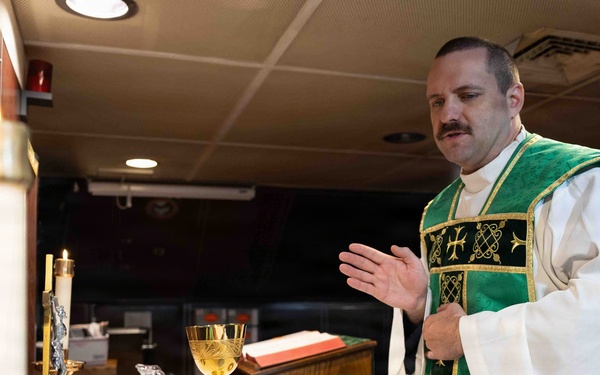 A Catholic priest wearing green liturgical vestments (chasuble) over a white alb is celebrating Mass at an altar. The altar is set with traditional Catholic Mass items including a chalice, candles, and an open missal. The setting appears to be a non-traditional chapel space, possibly aboard a military vessel or in a converted room.