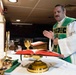 A Catholic priest wearing green liturgical vestments (chasuble) over a white alb is celebrating Mass at an altar. The altar is set with traditional Catholic Mass items including a chalice, candles, and an open missal. The setting appears to be a non-traditional chapel space, possibly aboard a military vessel or in a converted room.