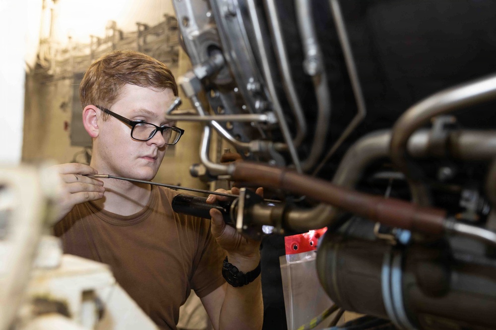 A young male technician wearing glasses and a brown t-shirt is performing maintenance or inspection work on a complex mechanical assembly, likely an aircraft engine or turbine. He is holding a tool, possibly a grease gun or inspection probe, and appears to be carefully examining or lubricating a component. The setting appears to be a maintenance bay or hangar environment.