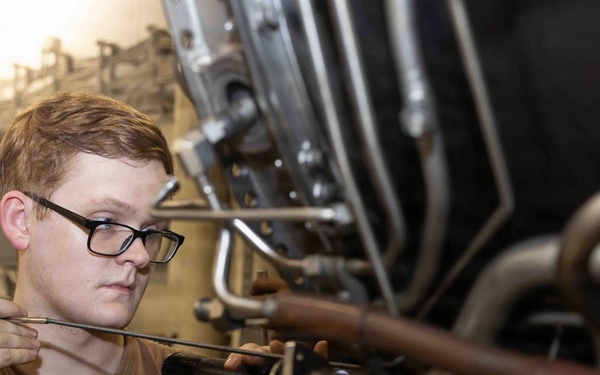 A young male technician wearing glasses and a brown t-shirt is performing maintenance or inspection work on a complex mechanical assembly, likely an aircraft engine or turbine. He is holding a tool, possibly a grease gun or inspection probe, and appears to be carefully examining or lubricating a component. The setting appears to be a maintenance bay or hangar environment.