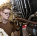 A young male technician wearing glasses and a brown t-shirt is performing maintenance or inspection work on a complex mechanical assembly, likely an aircraft engine or turbine. He is holding a tool, possibly a grease gun or inspection probe, and appears to be carefully examining or lubricating a component. The setting appears to be a maintenance bay or hangar environment.