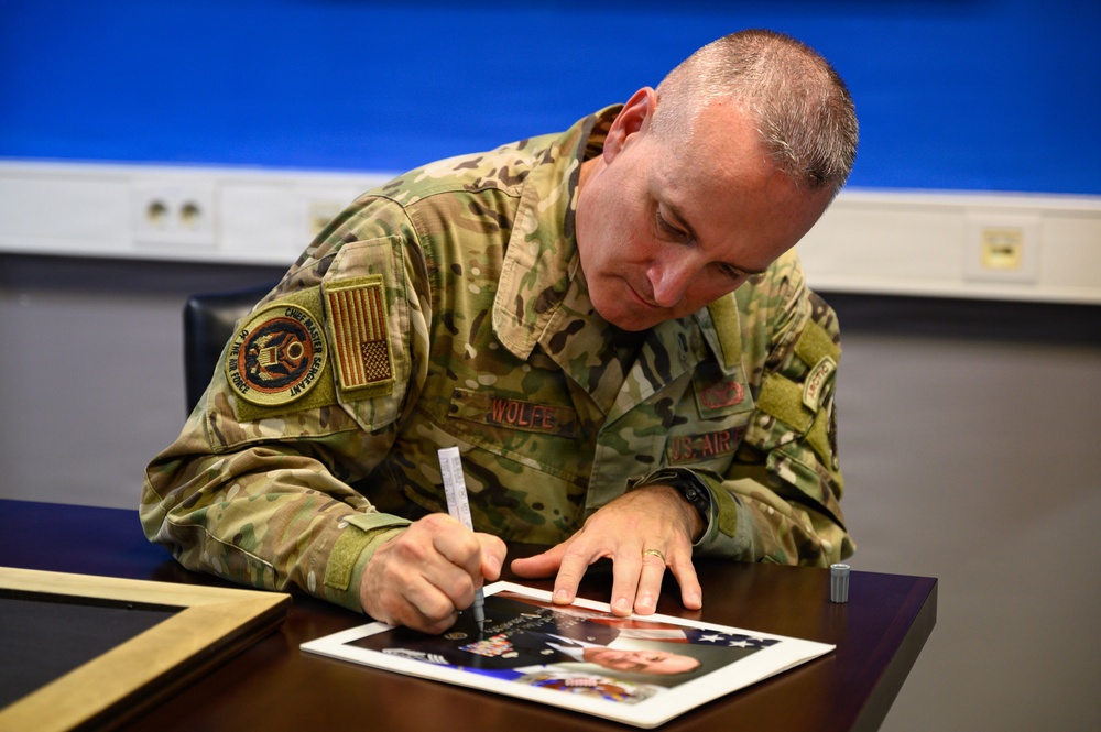 A U.S. Air Force Chief Master Sergeant named Wolfe is signing a photograph at a desk. He is wearing an OCP (Operational Camouflage Pattern) uniform with a name tape reading 'WOLFE' and a Chief Master Sergeant insignia patch. The setting appears to be an official military environment, likely during a formal or ceremonial occasion.