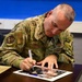 A U.S. Air Force Chief Master Sergeant named Wolfe is signing a photograph at a desk. He is wearing an OCP (Operational Camouflage Pattern) uniform with a name tape reading 'WOLFE' and a Chief Master Sergeant insignia patch. The setting appears to be an official military environment, likely during a formal or ceremonial occasion.