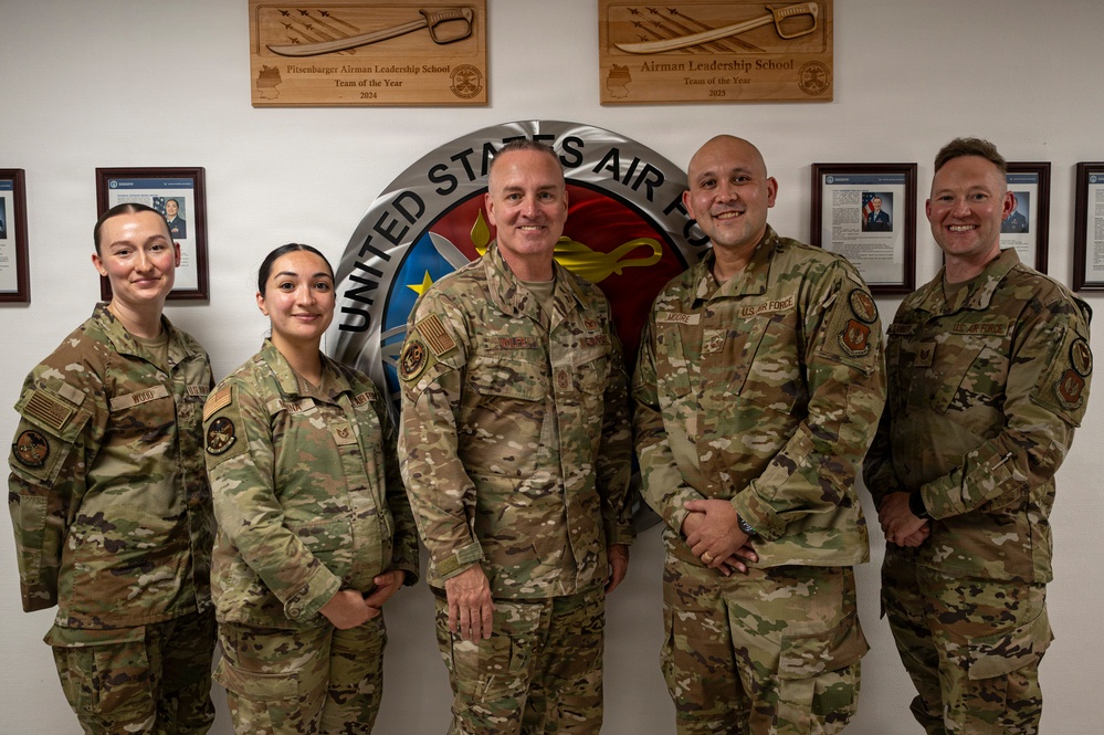 Five United States Air Force personnel in OCP (Operational Camouflage Pattern) uniforms pose for a group photo in front of a United States Air Force emblem/seal mounted on a wall. The setting appears to be an official Air Force facility, likely associated with Airman Leadership School based on the plaques visible on the wall. The group consists of two female and three male service members smiling for the camera.