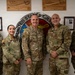 Five United States Air Force personnel in OCP (Operational Camouflage Pattern) uniforms pose for a group photo in front of a United States Air Force emblem/seal mounted on a wall. The setting appears to be an official Air Force facility, likely associated with Airman Leadership School based on the plaques visible on the wall. The group consists of two female and three male service members smiling for the camera.