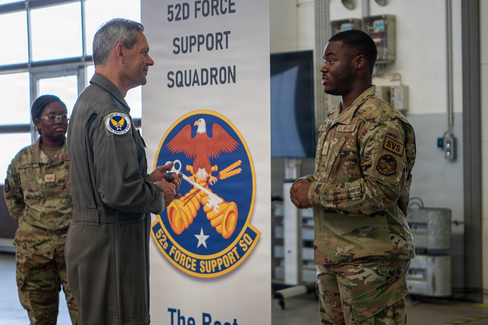 Two U.S. Air Force personnel are engaged in conversation in front of a 52D Force Support Squadron banner. A senior officer in a flight suit is speaking with a junior enlisted airman in OCP (Operational Camouflage Pattern) uniform, while a third service member stands in the background. The setting appears to be an indoor military facility or hangar.