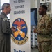 Two U.S. Air Force personnel are engaged in conversation in front of a 52D Force Support Squadron banner. A senior officer in a flight suit is speaking with a junior enlisted airman in OCP (Operational Camouflage Pattern) uniform, while a third service member stands in the background. The setting appears to be an indoor military facility or hangar.