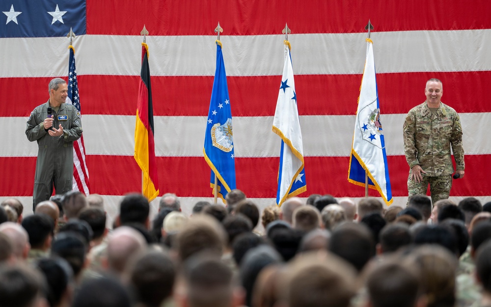 A military ceremony or all-hands event is taking place in what appears to be a large hangar or indoor venue, with two uniformed military personnel addressing a large assembled crowd. A massive American flag serves as a backdrop, flanked by several organizational and national flags including the German flag and US Air Force-related flags. The gathering appears to be a formal military event, possibly a change of command, briefing, or official address.