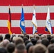 A military ceremony or all-hands event is taking place in what appears to be a large hangar or indoor venue, with two uniformed military personnel addressing a large assembled crowd. A massive American flag serves as a backdrop, flanked by several organizational and national flags including the German flag and US Air Force-related flags. The gathering appears to be a formal military event, possibly a change of command, briefing, or official address.