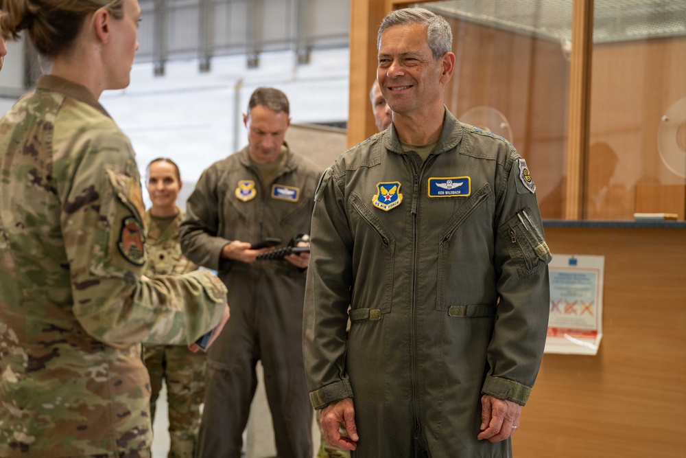 A U.S. Air Force officer wearing a flight suit with a name tag reading 'KEN WILSBACH' and a command patch is engaged in conversation with another service member in multicam uniform. The setting appears to be an indoor military facility, likely a hangar or operations building. Several other military personnel are visible in the background.