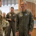 A U.S. Air Force officer wearing a flight suit with a name tag reading 'KEN WILSBACH' and a command patch is engaged in conversation with another service member in multicam uniform. The setting appears to be an indoor military facility, likely a hangar or operations building. Several other military personnel are visible in the background.