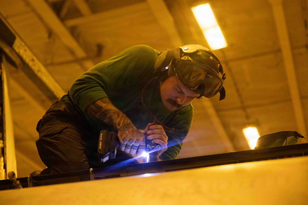 A heavily tattooed male worker is actively welding a metal surface inside an industrial facility. He is wearing a welding helmet pushed up on his head and is bent over his work, with a bright blue-white welding arc visible at the point of contact. The scene is illuminated by both artificial overhead lighting and the intense light of the welding process.
