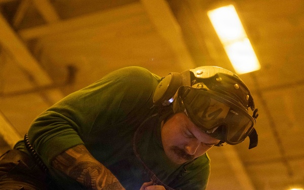 A heavily tattooed male worker is actively welding a metal surface inside an industrial facility. He is wearing a welding helmet pushed up on his head and is bent over his work, with a bright blue-white welding arc visible at the point of contact. The scene is illuminated by both artificial overhead lighting and the intense light of the welding process.