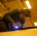 A heavily tattooed male worker is actively welding a metal surface inside an industrial facility. He is wearing a welding helmet pushed up on his head and is bent over his work, with a bright blue-white welding arc visible at the point of contact. The scene is illuminated by both artificial overhead lighting and the intense light of the welding process.