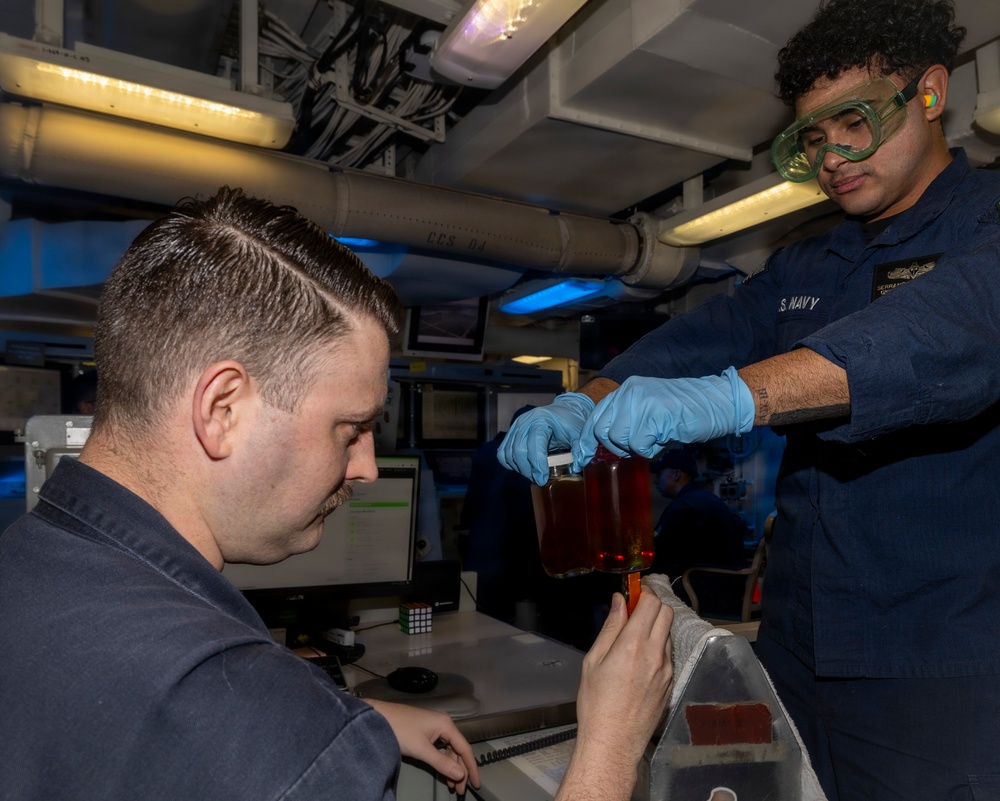 Two U.S. Navy personnel are conducting what appears to be a fluid or oil sampling/testing procedure aboard a naval vessel. One sailor wearing safety goggles and blue nitrile gloves is pouring or transferring a reddish-amber liquid from a container, while a seated sailor assists with the collection process. The setting is an interior compartment of a ship with visible piping, cable runs, and computer workstations.