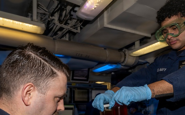 Two U.S. Navy personnel are conducting what appears to be a fluid or oil sampling/testing procedure aboard a naval vessel. One sailor wearing safety goggles and blue nitrile gloves is pouring or transferring a reddish-amber liquid from a container, while a seated sailor assists with the collection process. The setting is an interior compartment of a ship with visible piping, cable runs, and computer workstations.