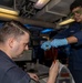 Two U.S. Navy personnel are conducting what appears to be a fluid or oil sampling/testing procedure aboard a naval vessel. One sailor wearing safety goggles and blue nitrile gloves is pouring or transferring a reddish-amber liquid from a container, while a seated sailor assists with the collection process. The setting is an interior compartment of a ship with visible piping, cable runs, and computer workstations.